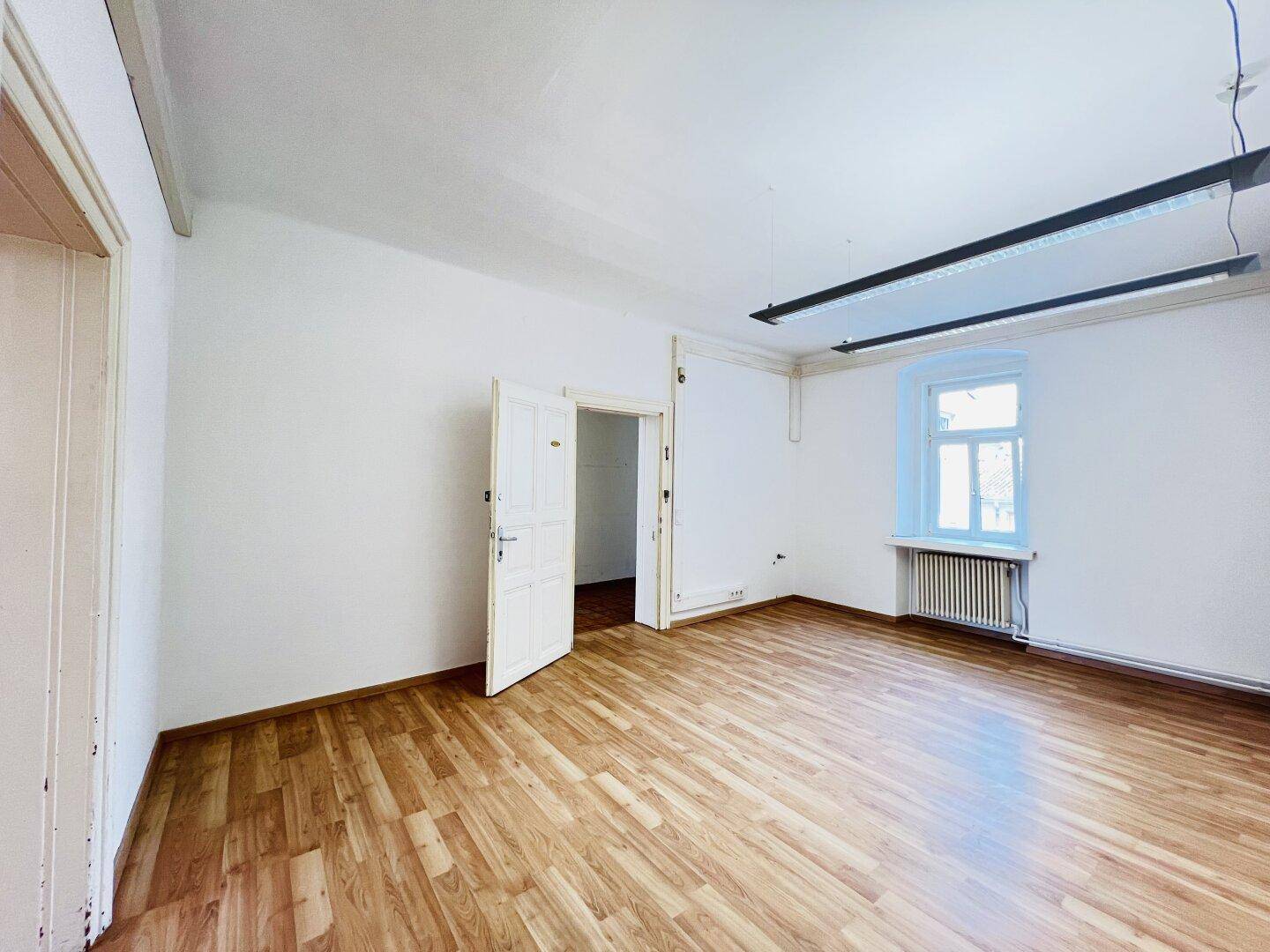 Heller Raum mit Laminatboden, einem Fenster und einer offenen Tür zu einem Nebenraum.