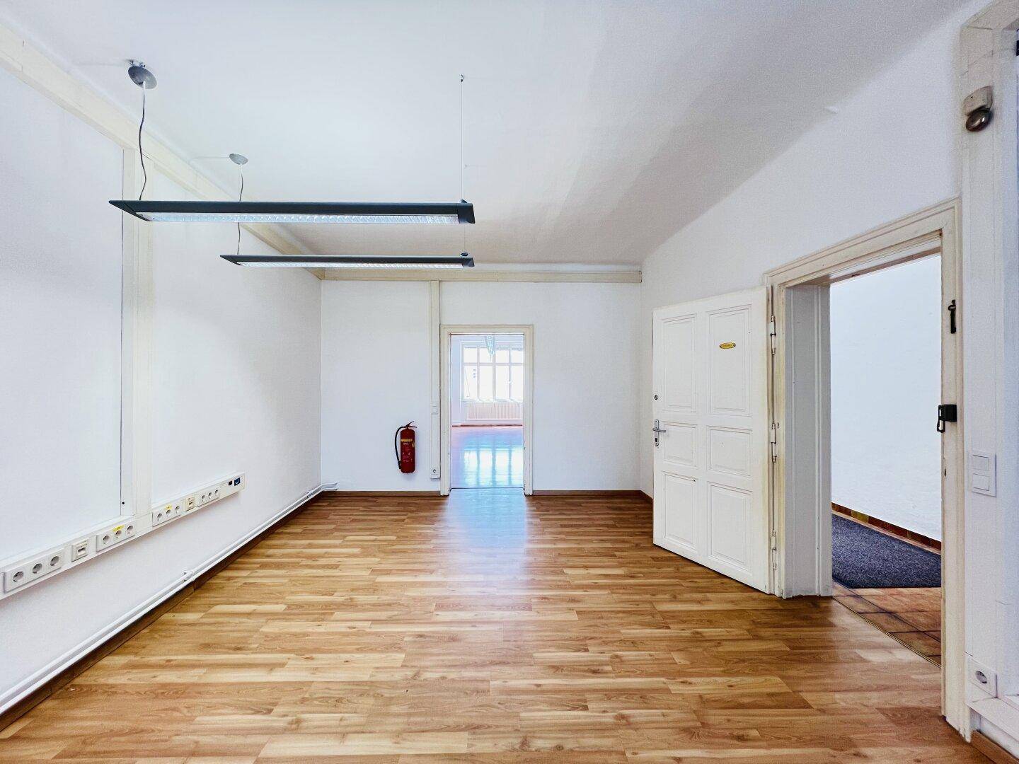 Heller Büroraum mit Holzboden, weißen Wänden und zwei modernen Hängelampen, mit Zugang zu weiteren Räumen.