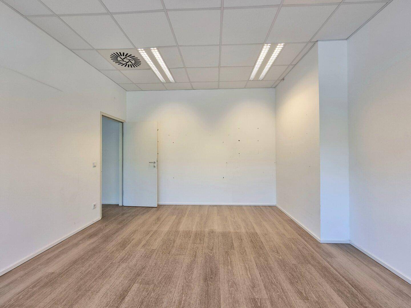 Leerer Büroraum mit Laminatboden, weißen Wänden, abgehängter Decke und einer weißen Tür.