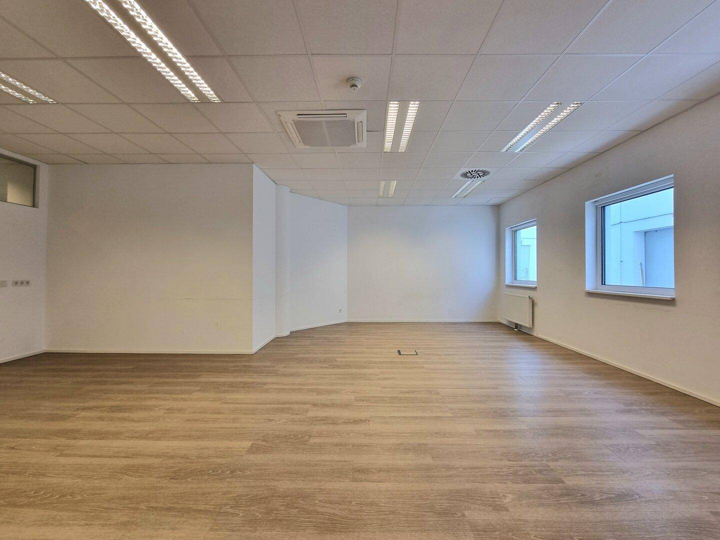 Geräumiger, leerer Büroraum mit Laminatboden, weißen Wänden, Deckenbeleuchtung und zwei Fenstern.