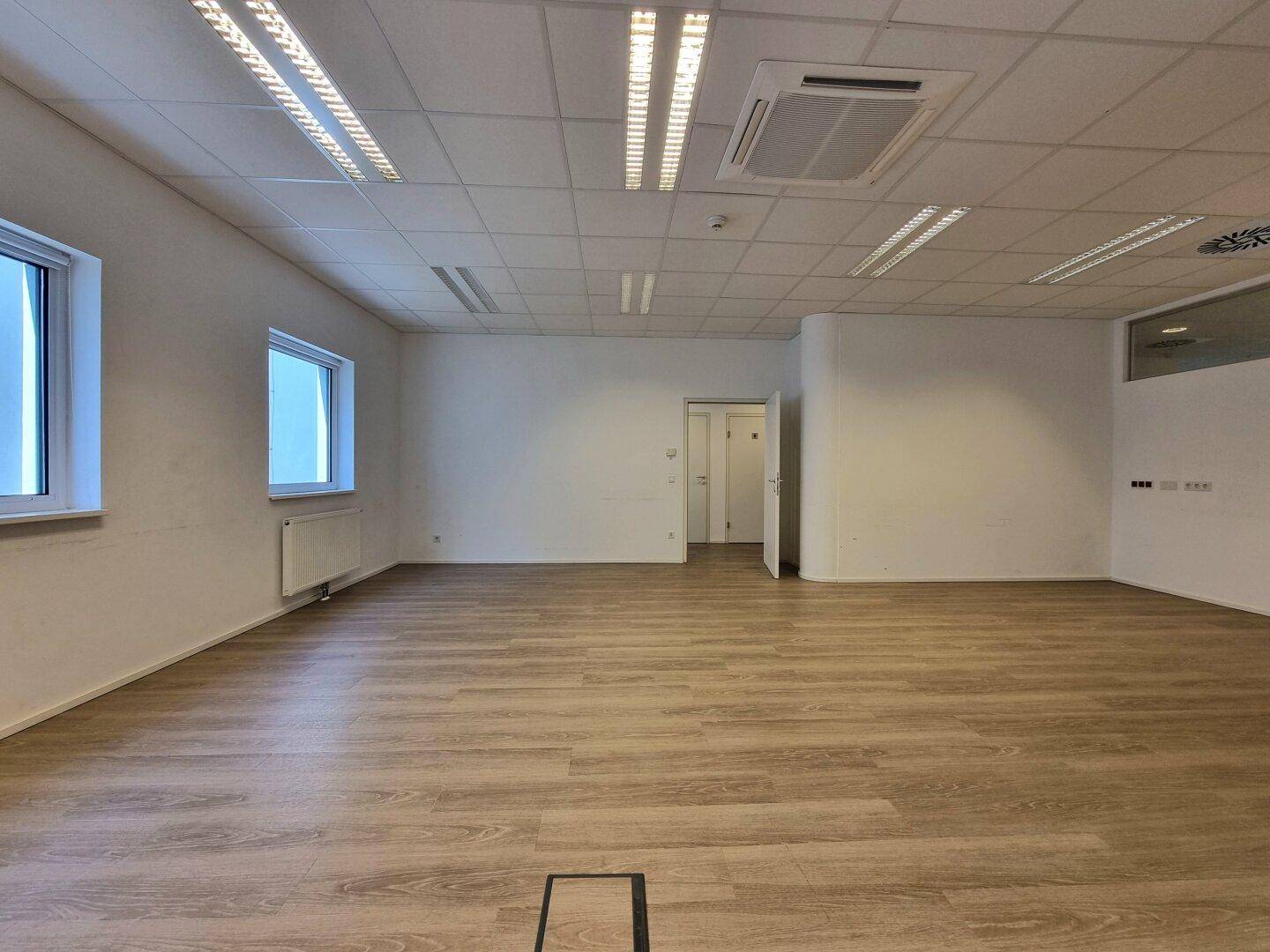 Heller Büroraum mit Laminatboden, weißen Wänden, abgehängter Decke und zwei Fenstern, die Tageslicht hereinlassen.