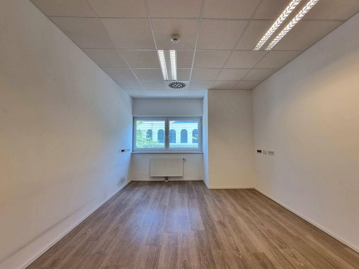 Helles, leeres Büro mit Fenster, Heizkörper und modernem Holzfußboden.