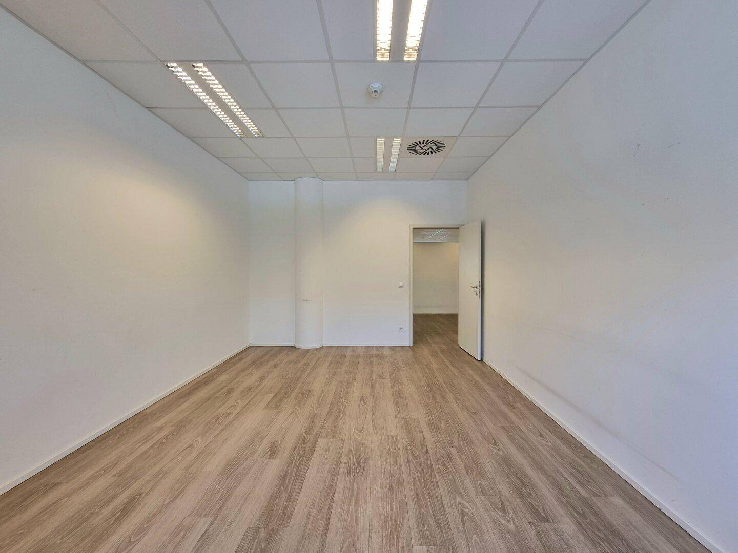 Leeres Büro mit holzbodenähnlichem Boden, weißen Wänden und einer Tür zu einem weiteren Raum.