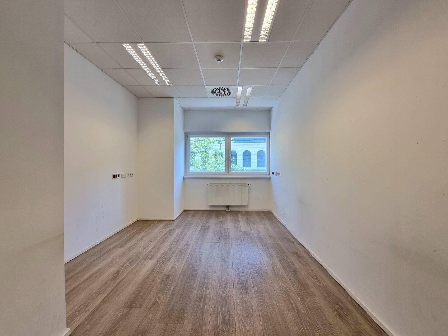 Kleinerer Büroraum mit Laminatboden, weißen Wänden, Deckenbeleuchtung und einem großen Fenster mit Grünblick.