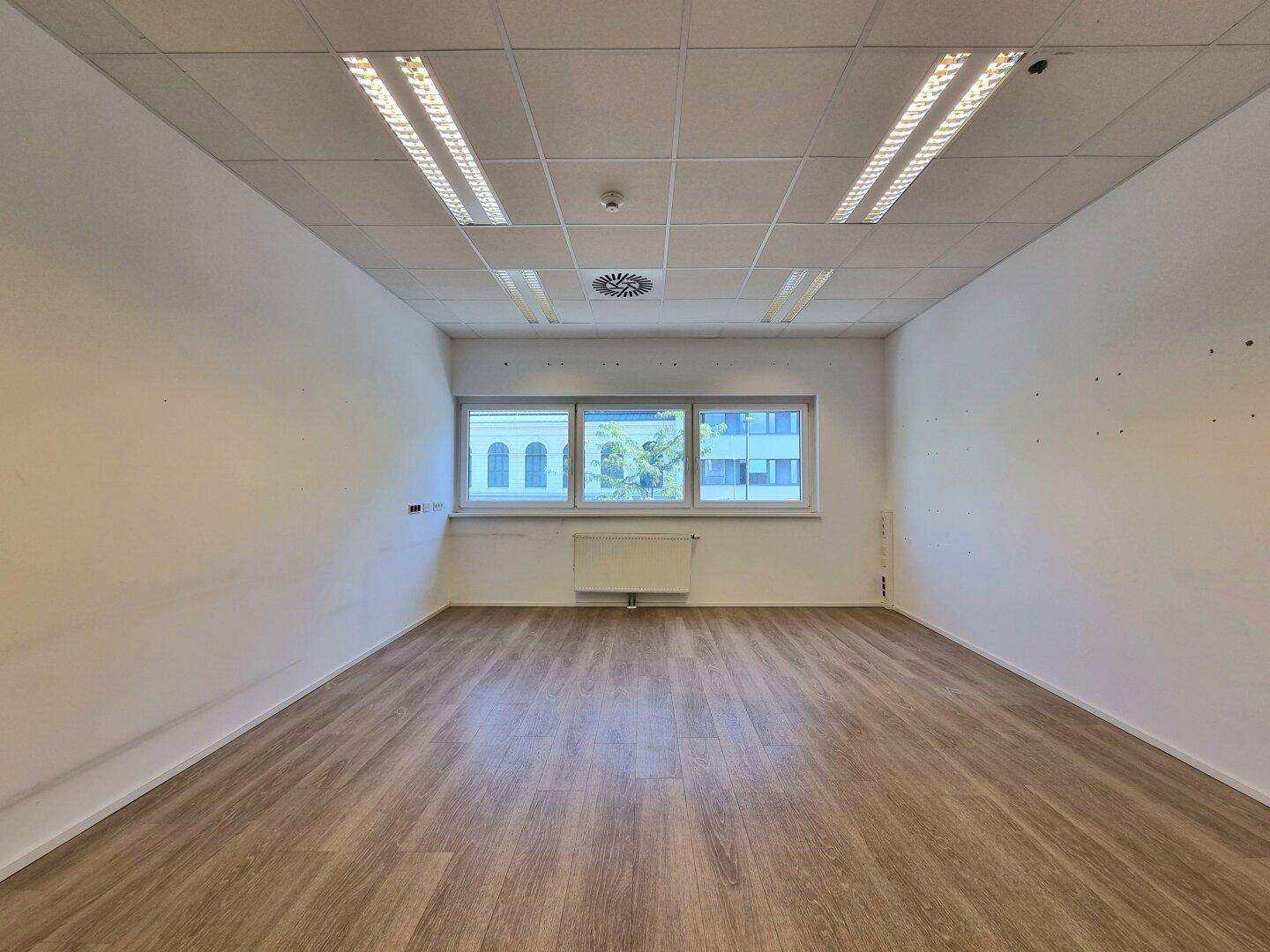 Geräumiges, helles Büro mit großem Fenster, Heizkörper und modernem Holzfußboden.