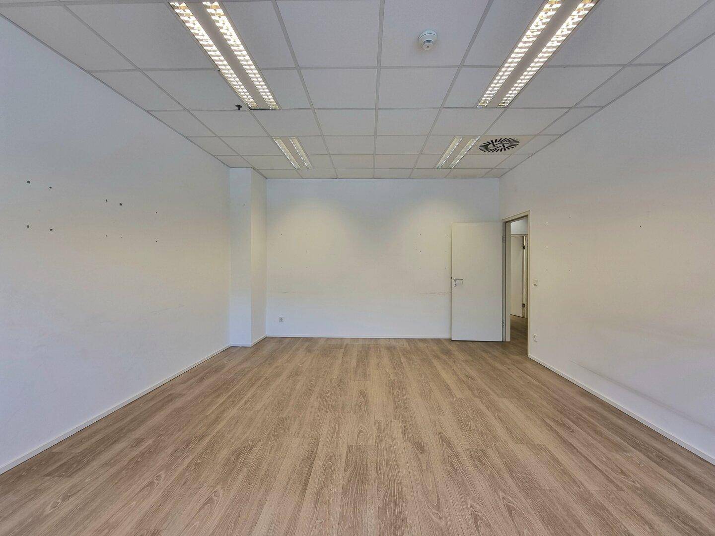 Geräumiger, leerer Büroraum mit Laminatboden, weißen Wänden, Deckenbeleuchtung und einer Tür.