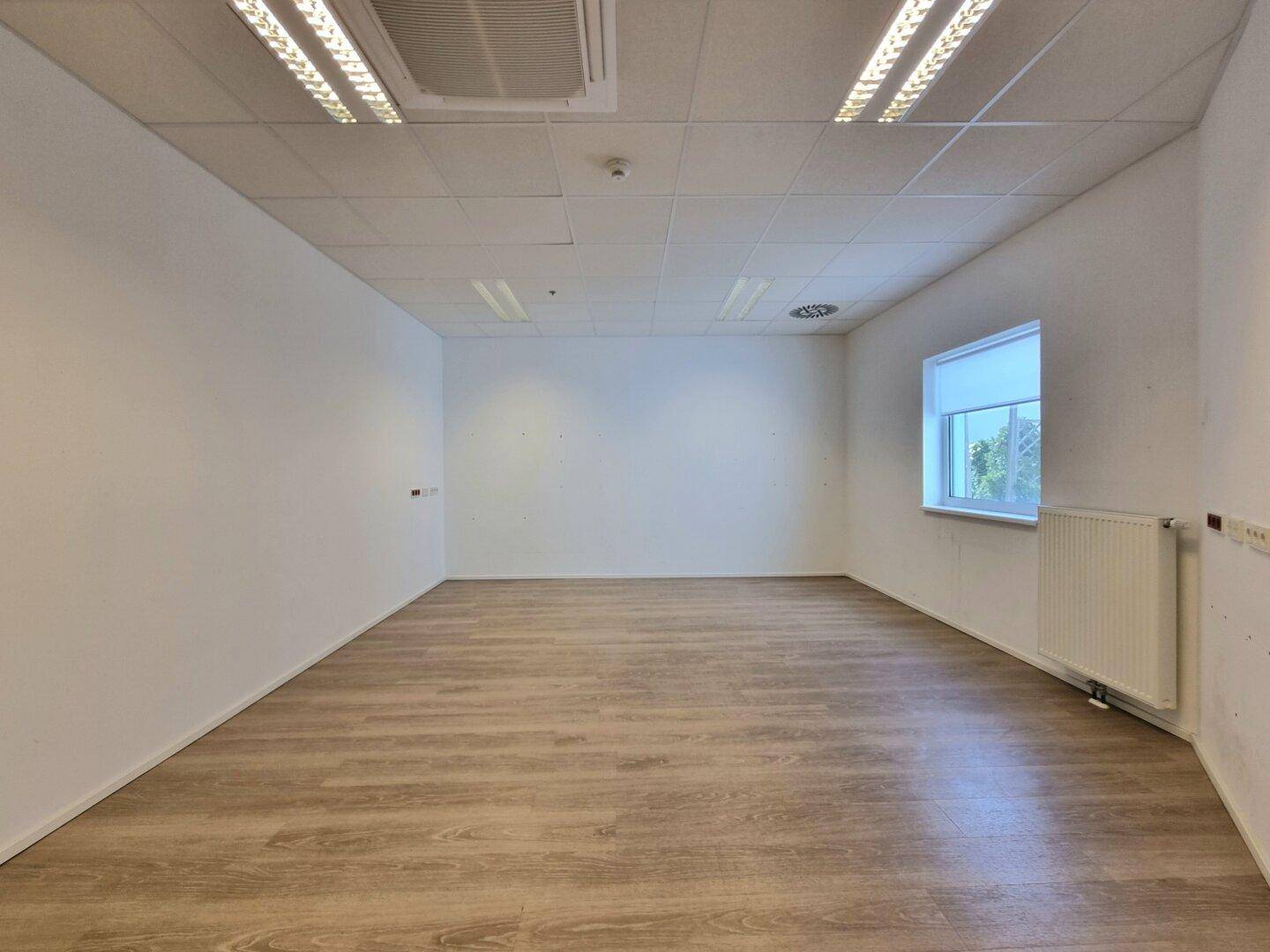 Heller Büroraum mit Laminatboden, weißen Wänden, abgehängter Decke, Klimaanlage und einem Fenster.