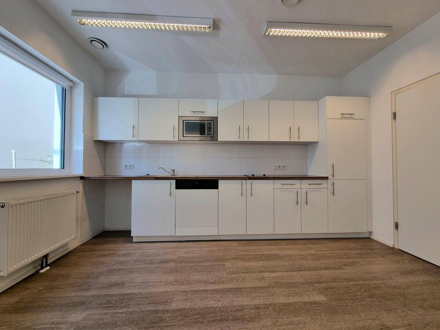 Moderne Küchenzeile mit weißen Schränken, Holzarbeitsplatte, Spülmaschine und Mikrowelle, neben einem Fenster.