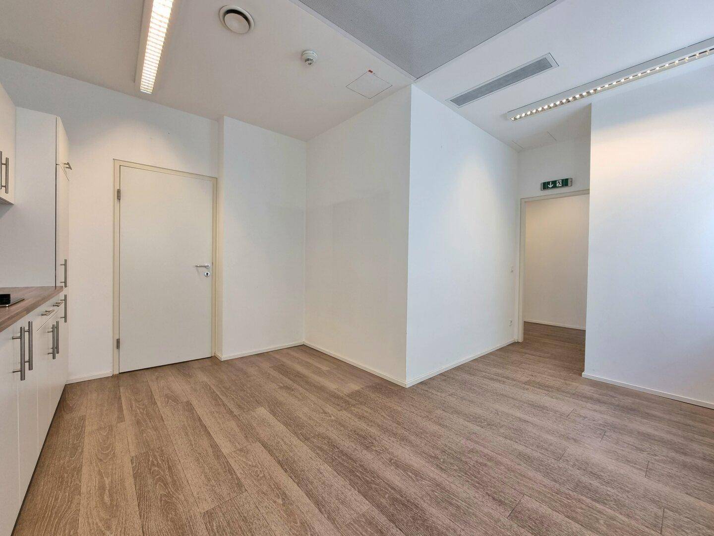 Büroraum mit kleiner Küchenzeile, hellem Holzfußboden und weißen Wänden.