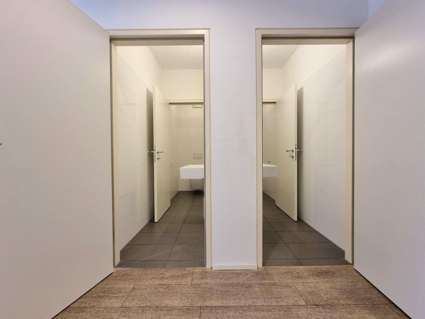 Zwei separate Toilettenräume mit weißen Fliesenwänden und dunklen Fliesenböden, jeweils mit Waschbecken.