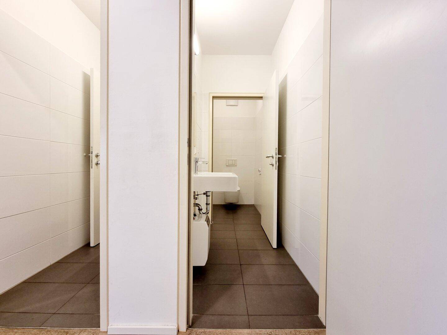 Heller Toilettenraum mit weißen Fliesenwänden, dunklem Fliesenboden, Waschbecken und Spiegel.