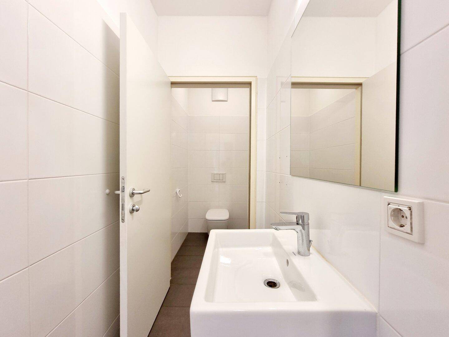 Detailansicht eines Waschbeckens mit Spiegel in einem gefliesten Toilettenraum, mit Toilette im Hintergrund.