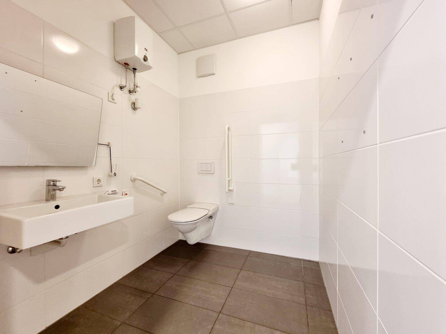 Barrierefreier Toilettenraum mit weißen Fliesen, dunklem Boden, Wand-WC und Haltegriffen für Zugänglichkeit.