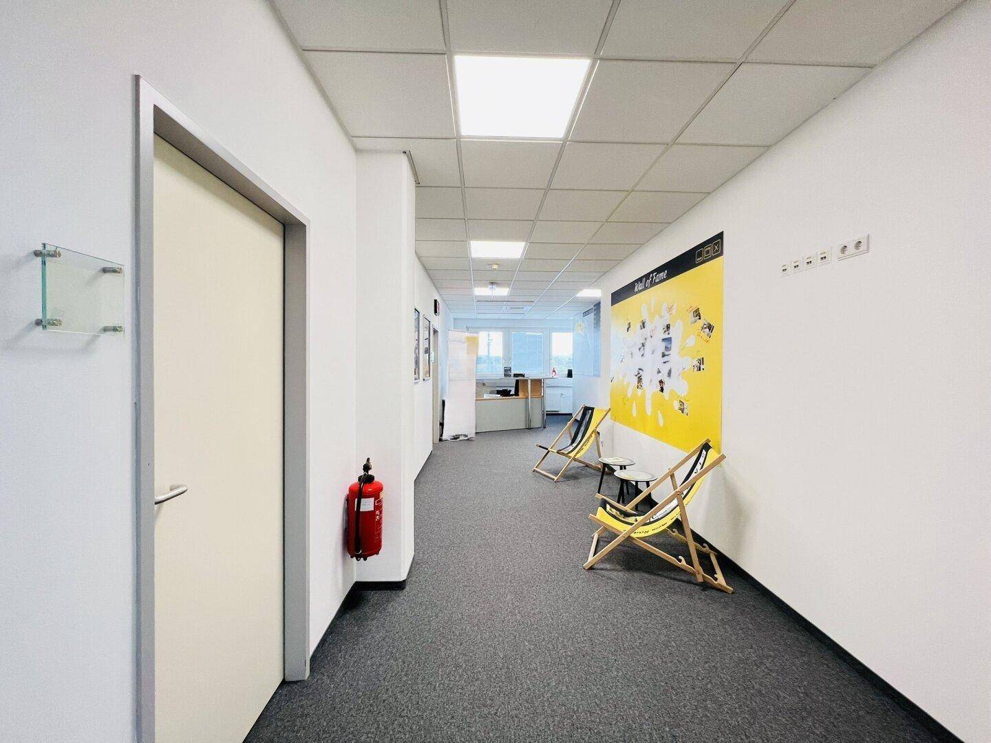 Heller Bürogang mit Teppichboden, Deckenbeleuchtung und Wanddekoration, führt zu weiteren Büroräumen.