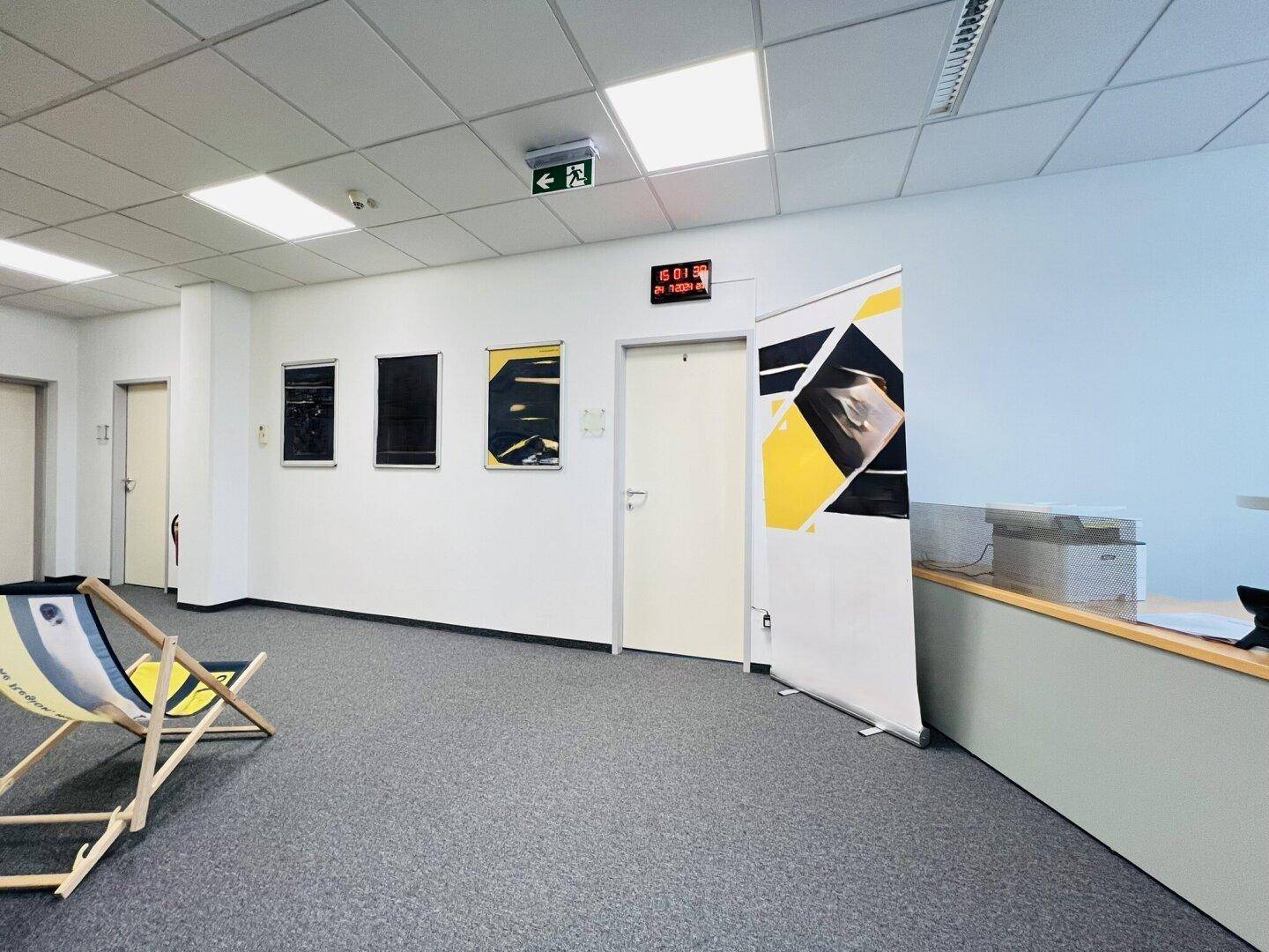 Bürogang mit Teppichboden, Deckenbeleuchtung und Wanddekoration, führt zu weiteren Büroräumen.