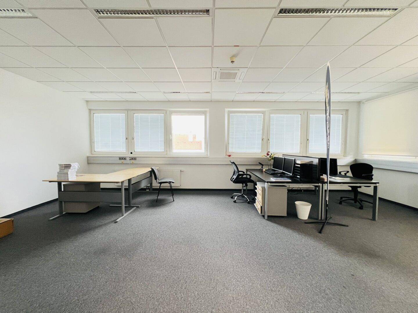 Großes, helles Büro mit Teppichboden, vielen Fenstern und mehreren leeren Schreibtischen.