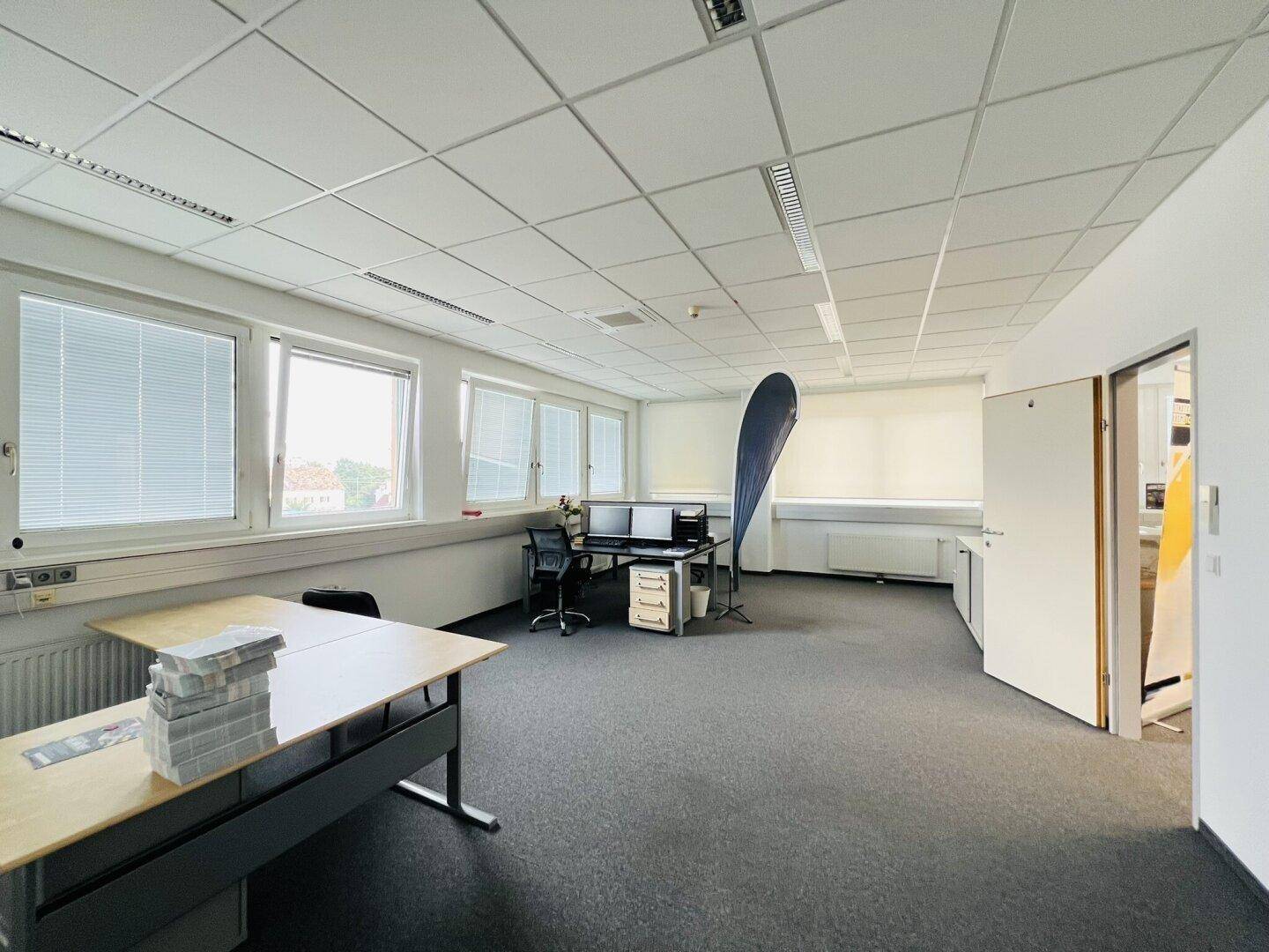 Geräumiges Büro mit Teppichboden, großen Fenstern und mehreren Arbeitsplätzen, gut beleuchtet.