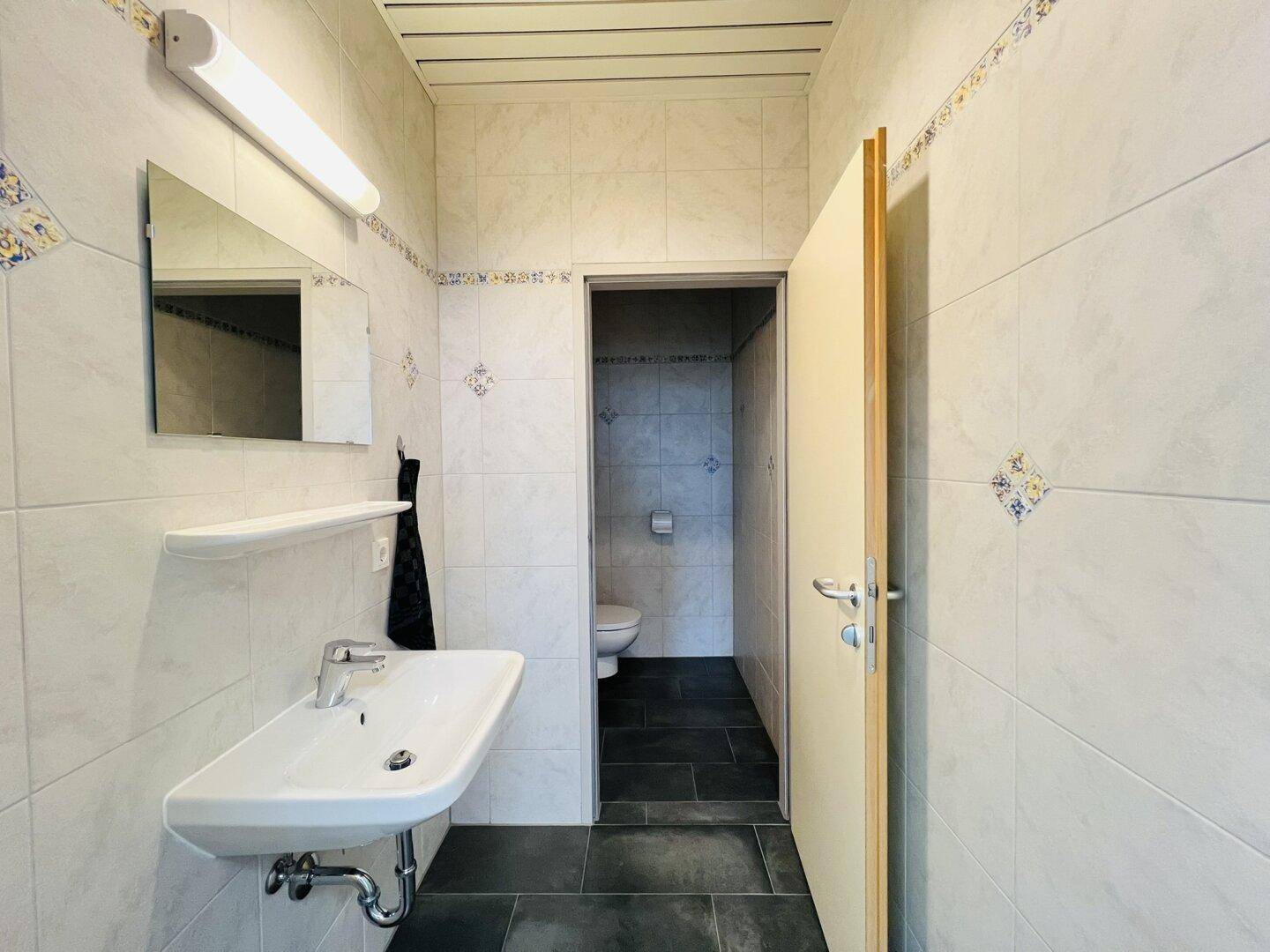 Gefliester Toilettenraum mit Waschbecken, Spiegel und separatem WC-Bereich, in gutem Zustand.