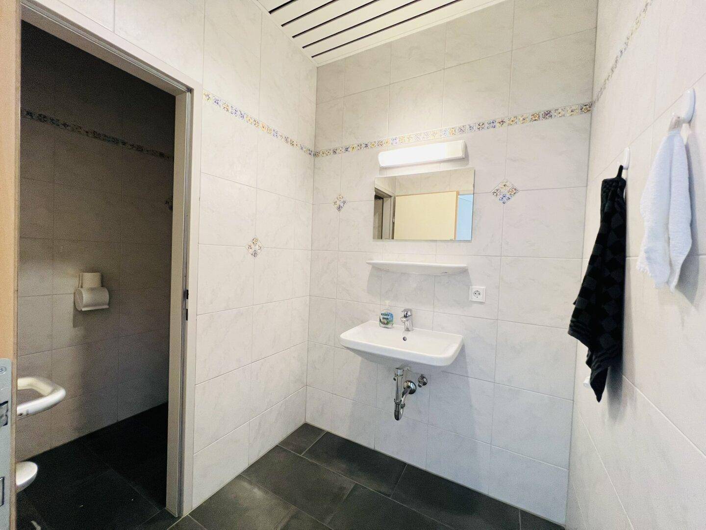 Gefliester Toilettenraum mit Waschbecken, Spiegel und separatem WC-Bereich, in gutem Zustand.