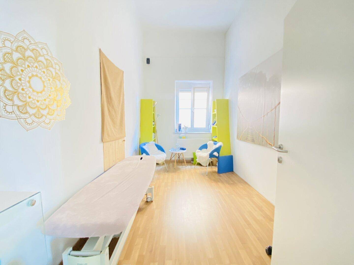 Ruhiger Therapieraum mit Liege, Holzboden, einem Fenster und dekorativer Wandkunst, ideal für Behandlungen.
