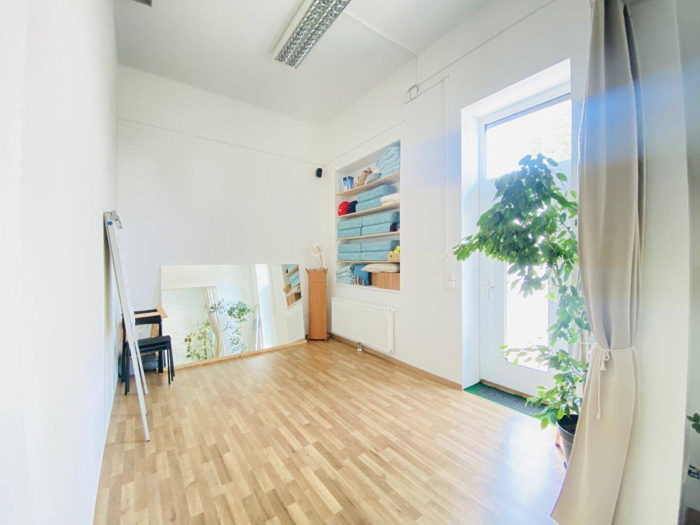 Heller Studiobereich mit Holzboden, großem Fenster zur Außenseite, Wandspiegel und praktischen Regalen.