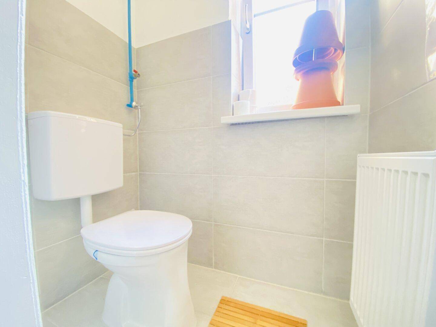 Helles Gäste-WC mit Toilette, Heizkörper und Fenster, komplett gefliest in modernem Grau.