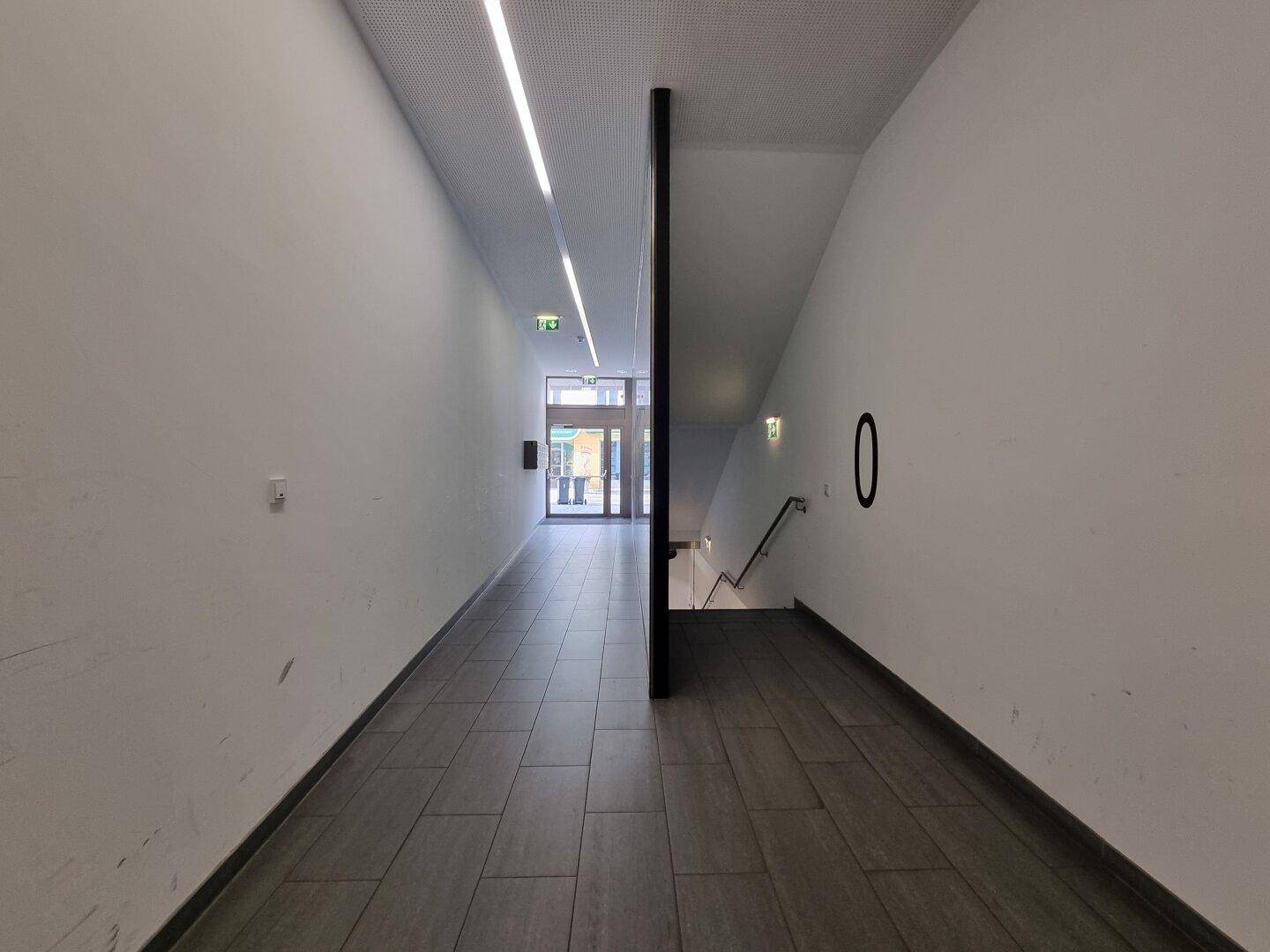 Langer, heller Flur mit Fliesenboden, der zu einem Eingangsbereich und einer Treppe im Gebäude führt.
