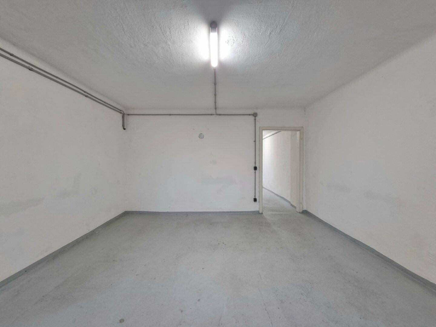 Geräumiger, leerer Kellerraum mit Betonboden, weißen Wänden und offenem Durchgang zu einem weiteren Bereich.