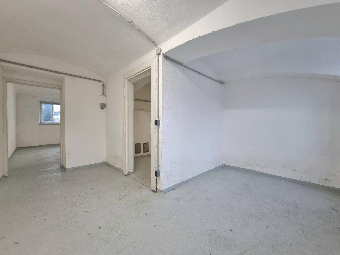 Mehrere leere Kellerräume mit Betonboden, weißen Wänden, sichtbaren Rohrleitungen und einem gewölbten Deckenbereich.