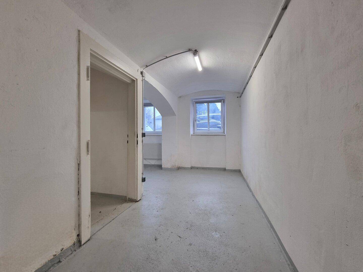 Leerer Kellerraum mit Betonboden, weißen Wänden, offenem Durchgang und zwei kleinen Fenstern für Belüftung.