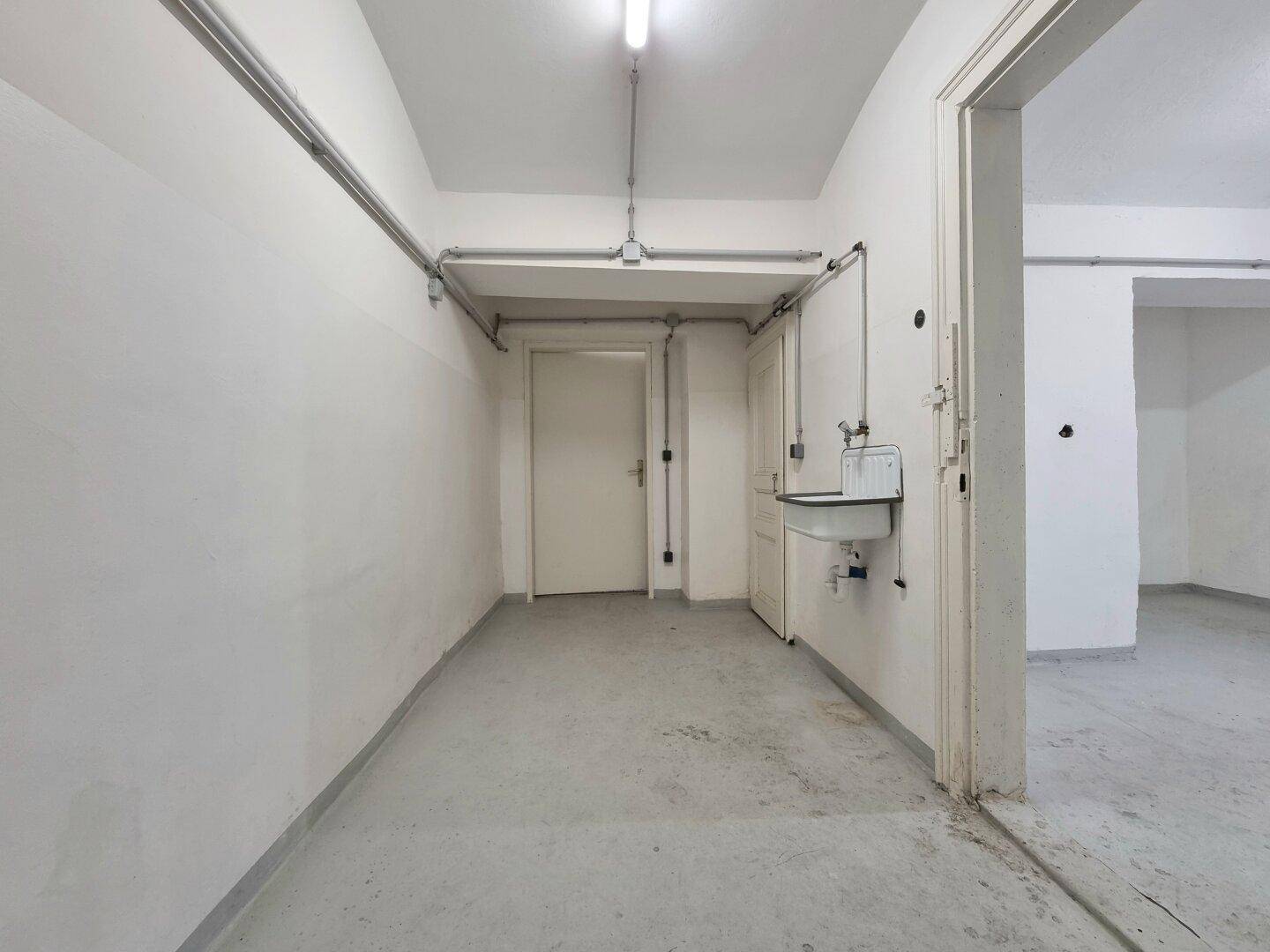Funktionaler Kellerraum mit Betonboden, Waschbecken und Zugang zu weiteren Räumen, ideal als Waschküche.