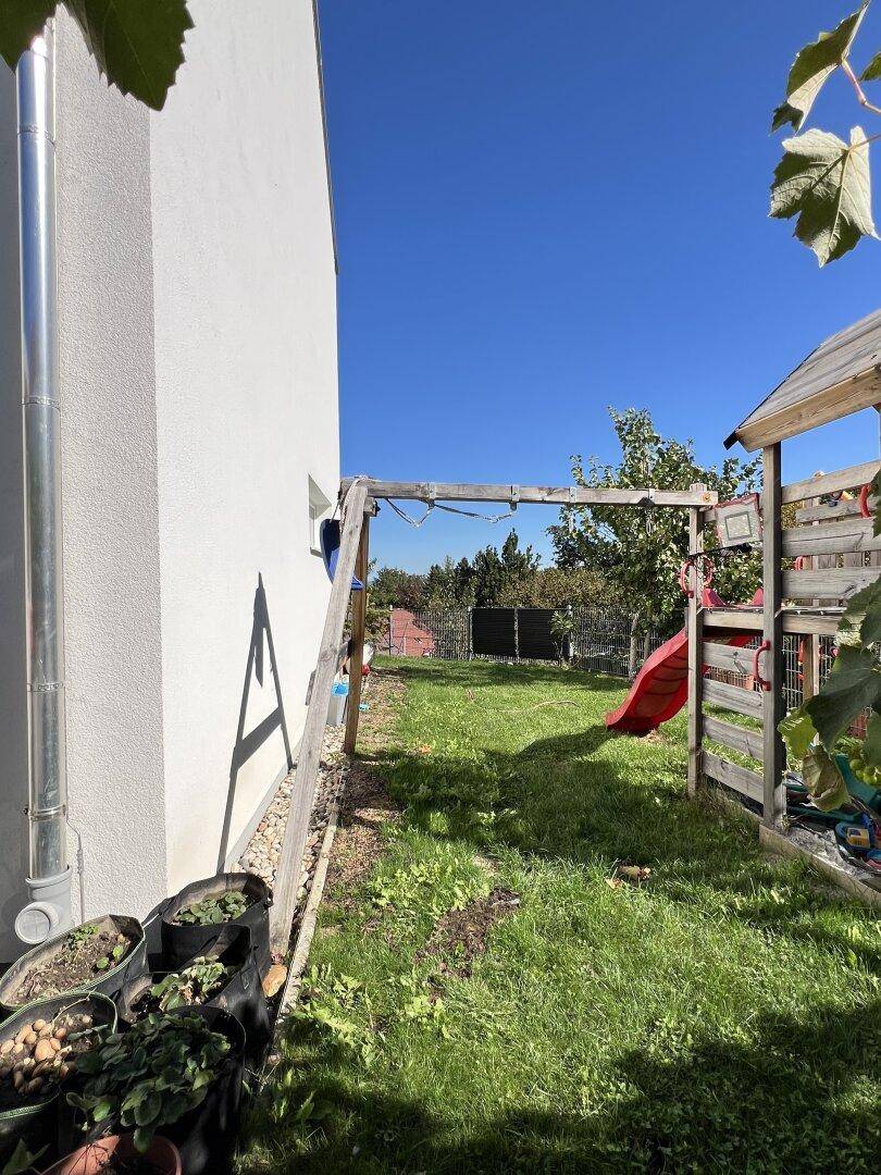 Seitlicher Gartenbereich mit Schaukel und Rutsche, umgeben von Grün und einem weißen Gebäude.