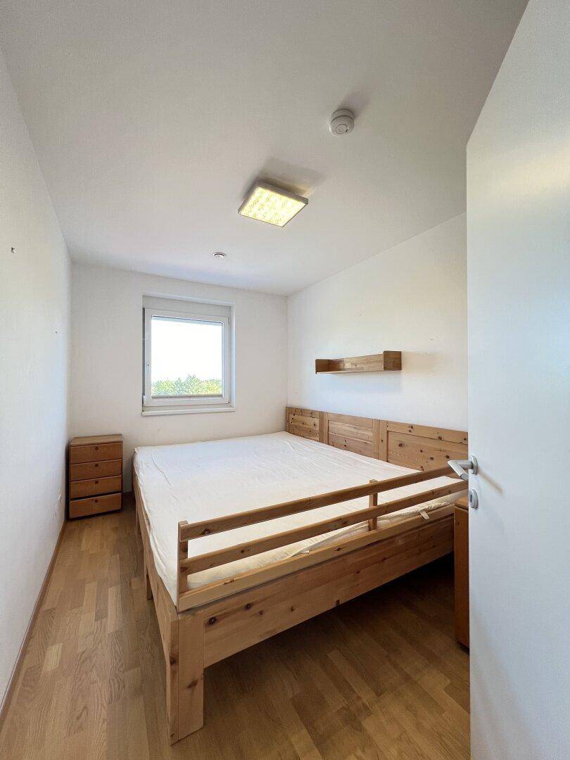 Schlafzimmer mit Holzbett, Nachttisch und Fenster, das viel Tageslicht hereinlässt.