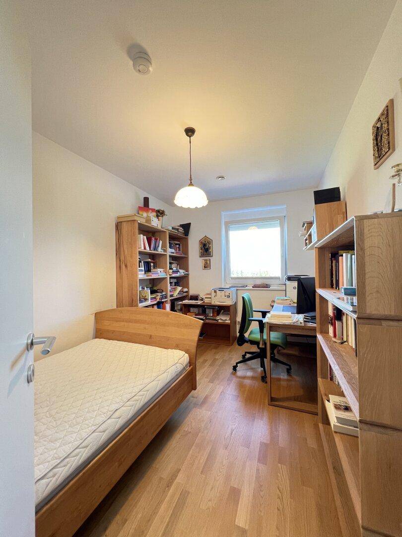 Helles Arbeitszimmer mit Einzelbett, Bücherregalen und Schreibtisch am Fenster.