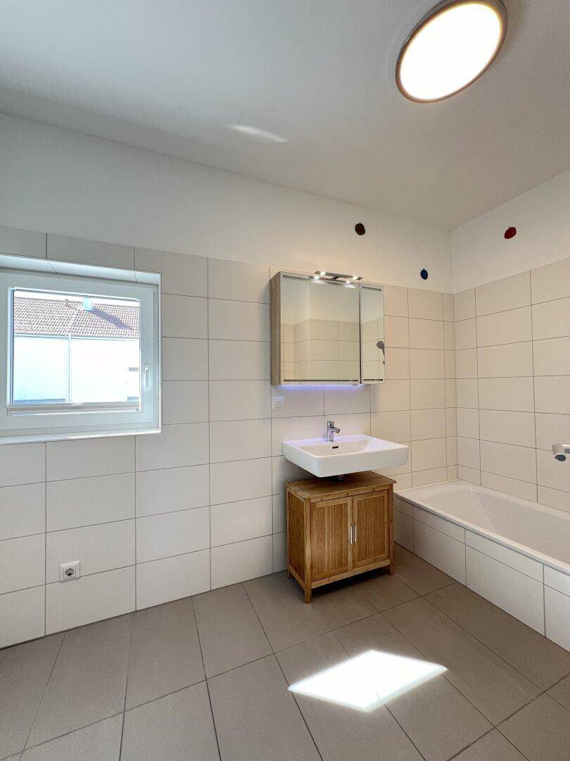 Helles Badezimmer mit Badewanne, Waschtisch, Spiegelschrank und Fenster für Tageslicht.