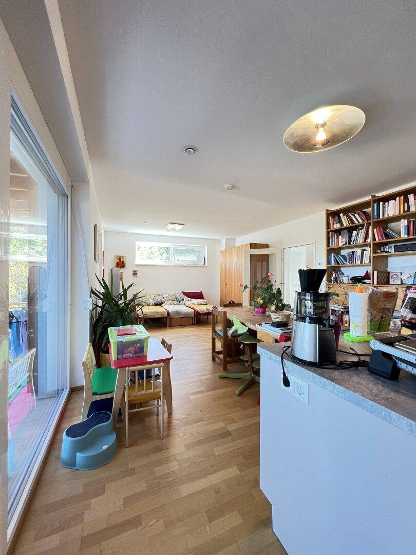 Offener Wohn-Essbereich mit Küche, Holzboden und großen Fenstern zum Außenbereich.