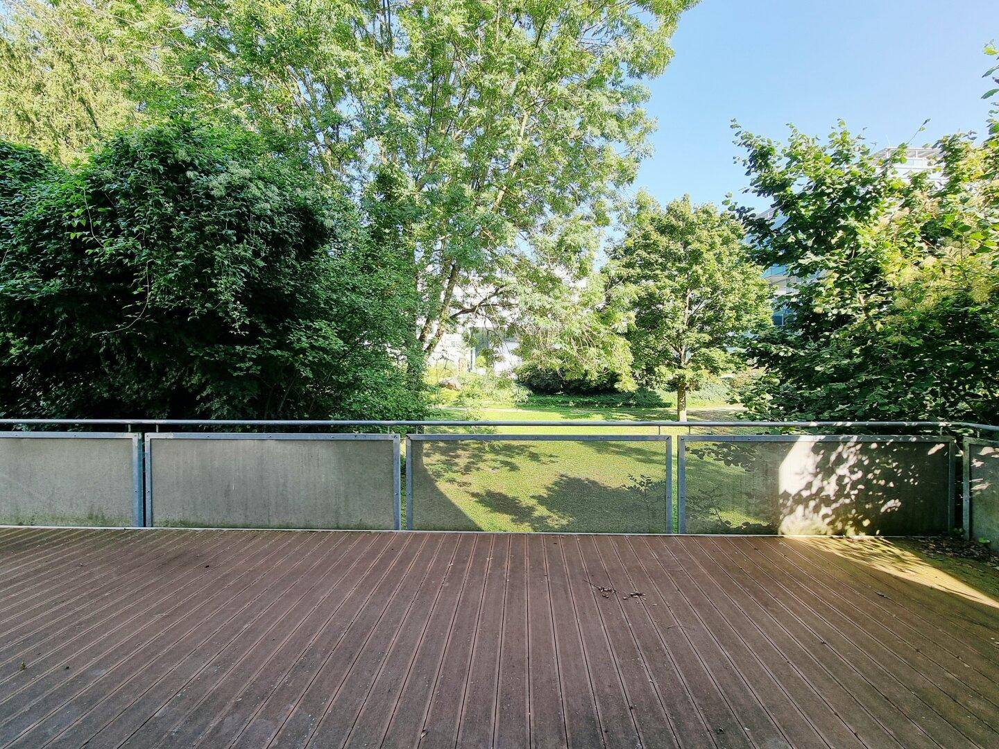 Weitläufige Terrasse mit Holzboden und Blick auf eine grüne Parklandschaft an einem sonnigen Tag.