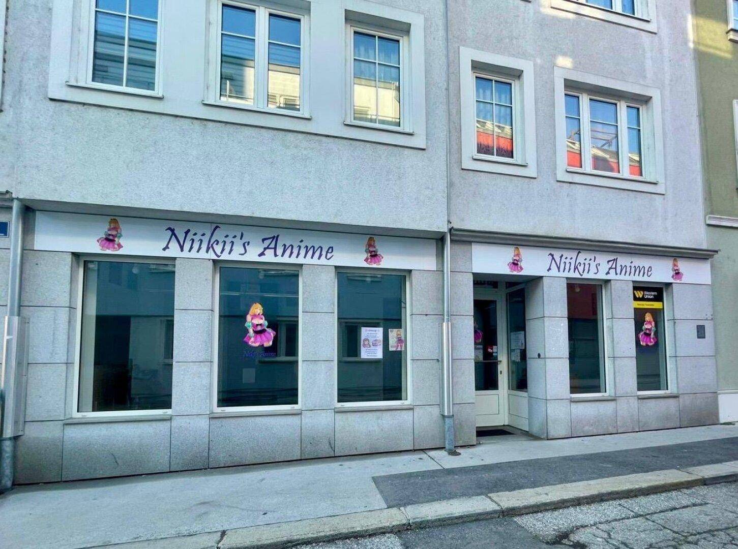Fassade eines Gebäudes mit einem Geschäft im Erdgeschoss und Wohnungen darüber, gekennzeichnet durch große Schaufenster und eine helle Putzfassade.