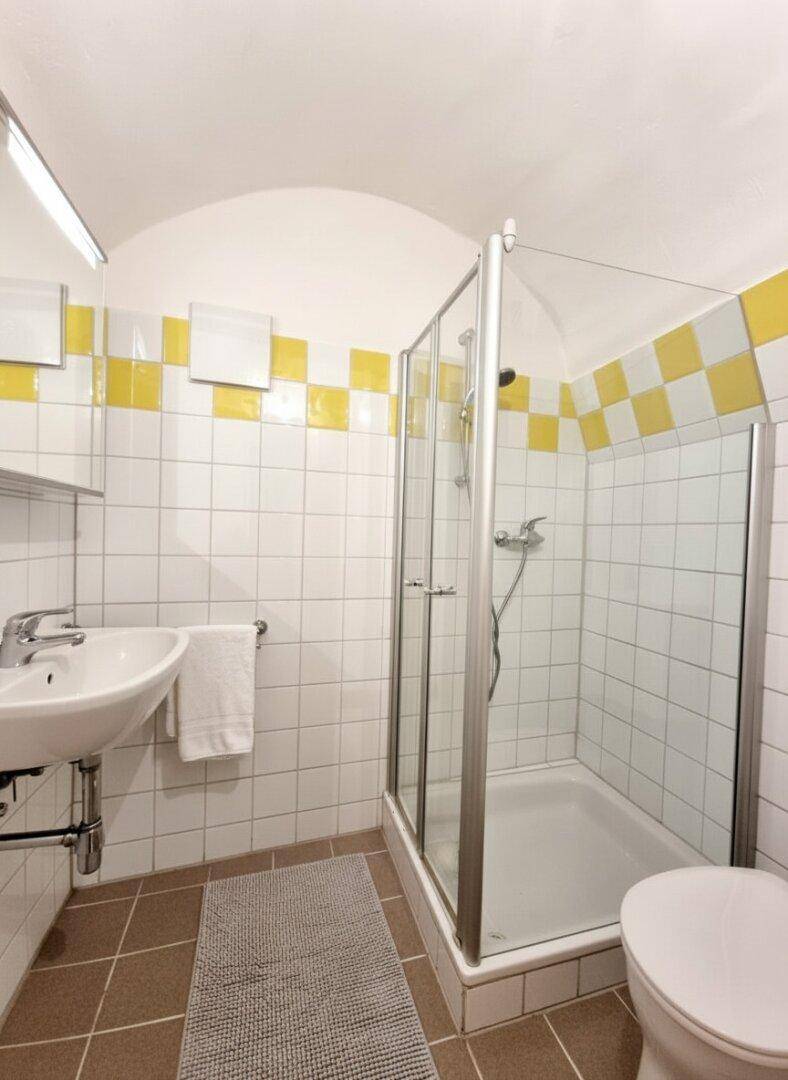 Badezimmer mit Duschkabine, Waschbecken und gelb-weißen Fliesen.