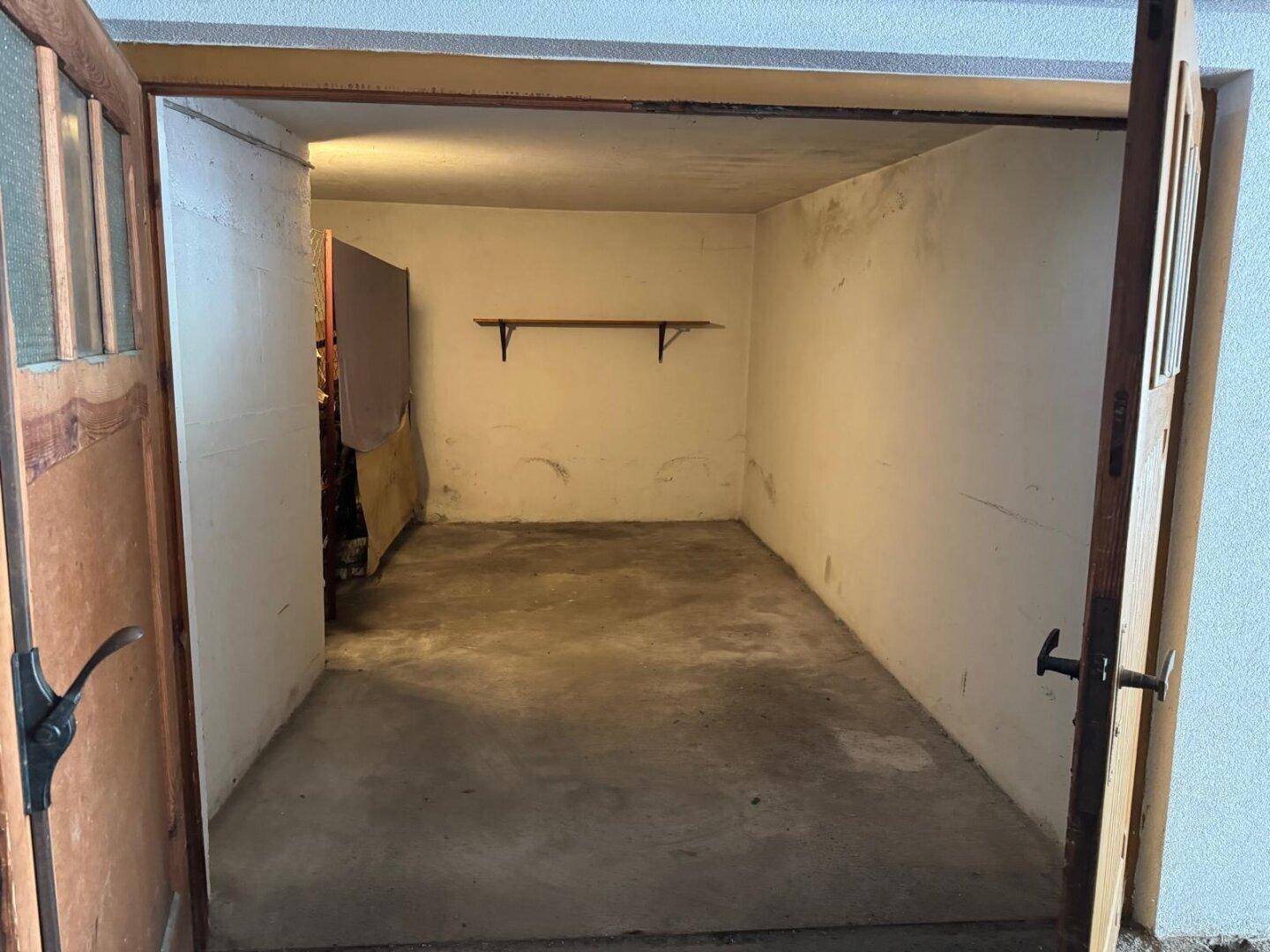 Leere Garage mit geöffneten Holztüren, Betonboden und einer einfachen Ablagefläche an der Wand.