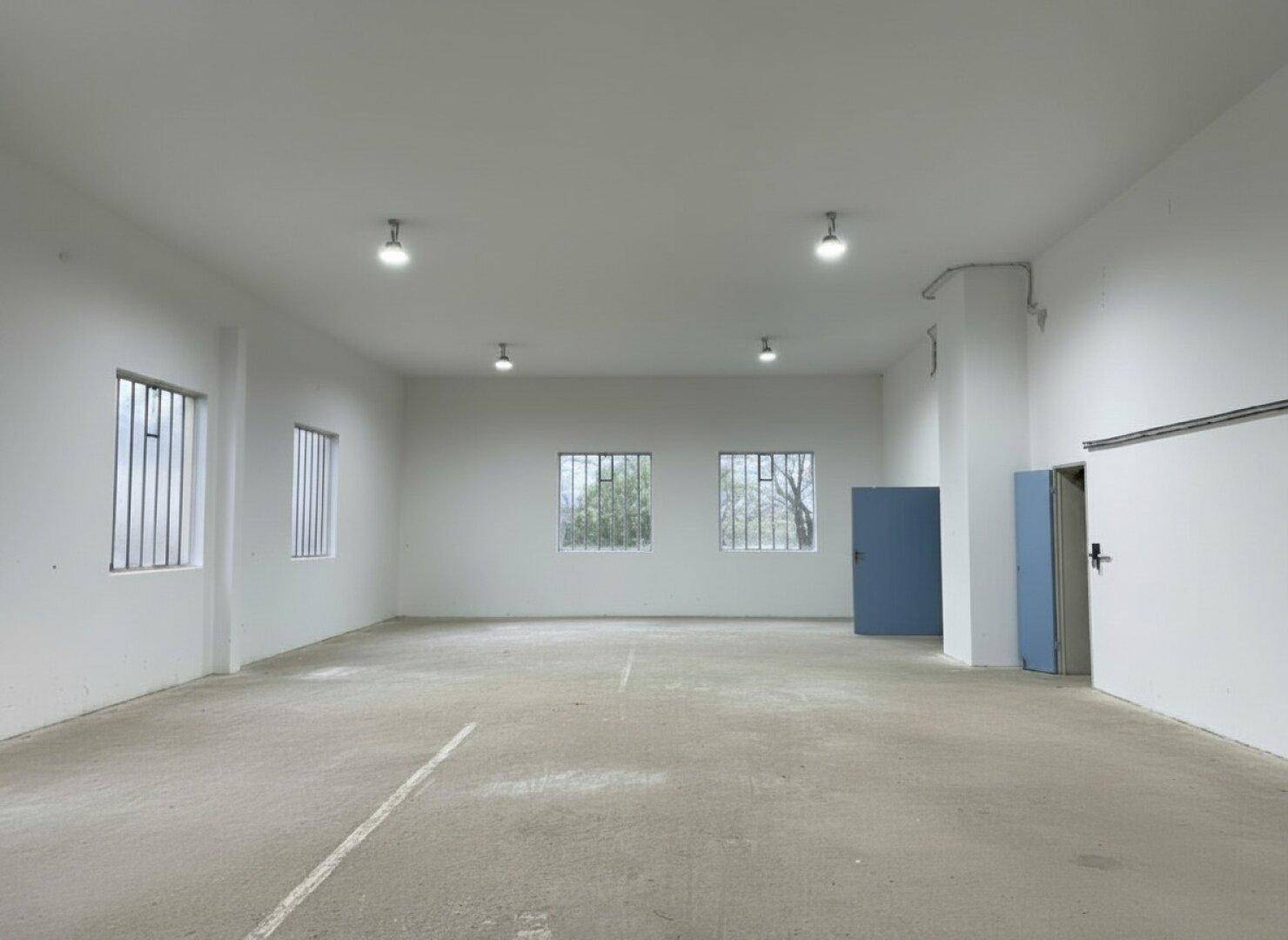 Helle, leere Lagerhalle mit mehreren Fenstern, Betonboden und einer blauen Tür.
