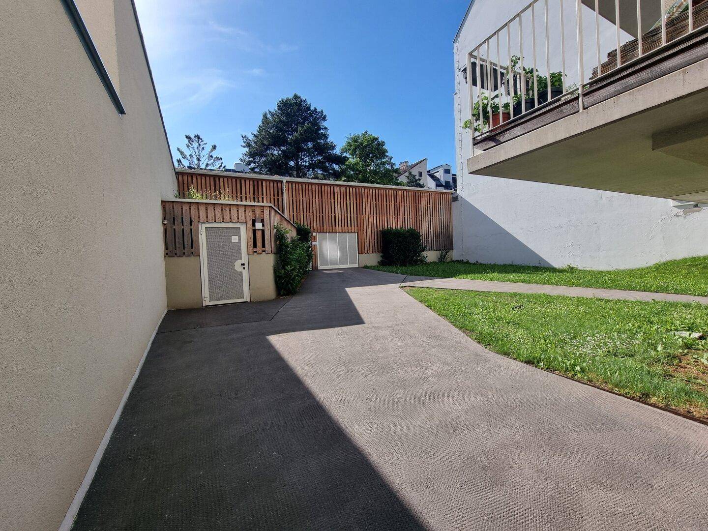 Außenbereich mit gepflastertem Weg, Rasenfläche und einem Balkon im Hintergrund, umgeben von Holzzäunen.