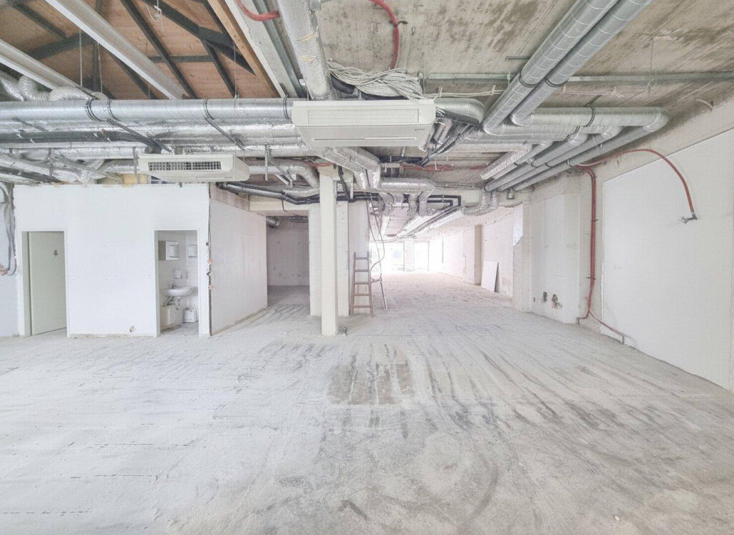 Weitläufiger Gewerberaum im Rohbau mit freiliegenden Lüftungsrohren und Installationen an der Decke sowie einem Teilausbau für Sanitäranlagen.