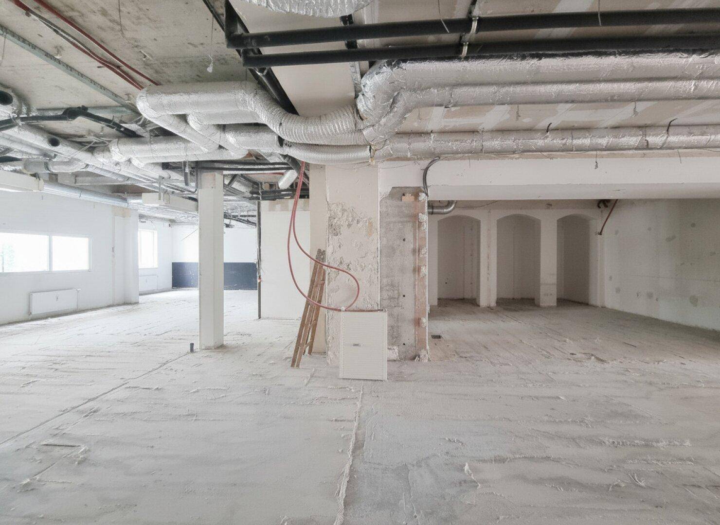 Großzügiger Gewerberaum im Rohbau mit sichtbaren Deckeninstallationen und unfertigen Wänden, durch Fenster fällt Tageslicht ein.