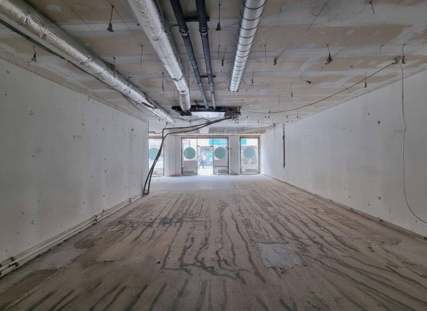 Langer Gewerberaum im Rohbau mit freiliegenden Deckeninstallationen und großen runden Fenstern am Ende des Raumes.