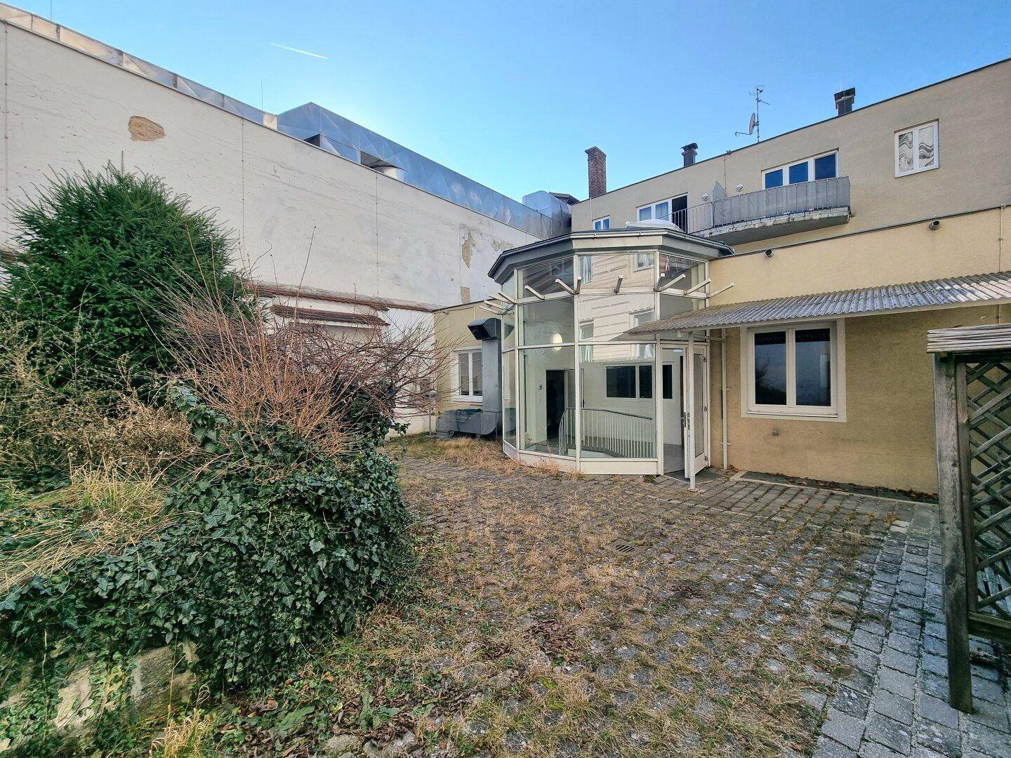 Außenansicht eines Gebäudes mit einem verglasten Eingangsbereich und einem ungepflegten Garten im Vordergrund.