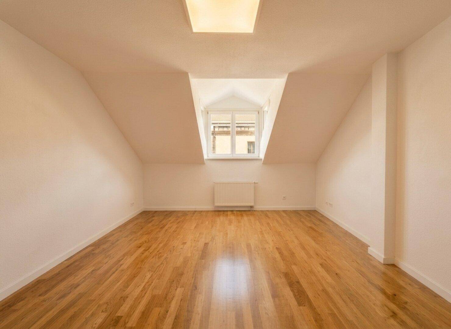 Helles Zimmer im Dachgeschoss mit schrägen Wänden, Holzfußboden und Fenster.