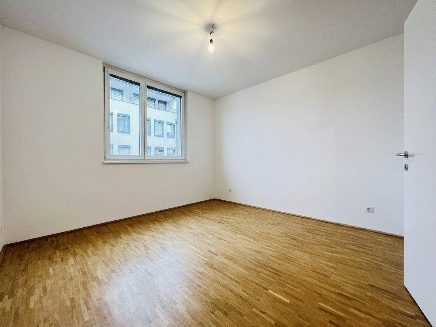 Leeres Zimmer mit hellem Parkettboden und einem Fenster mit Blick auf die umliegenden Gebäude.