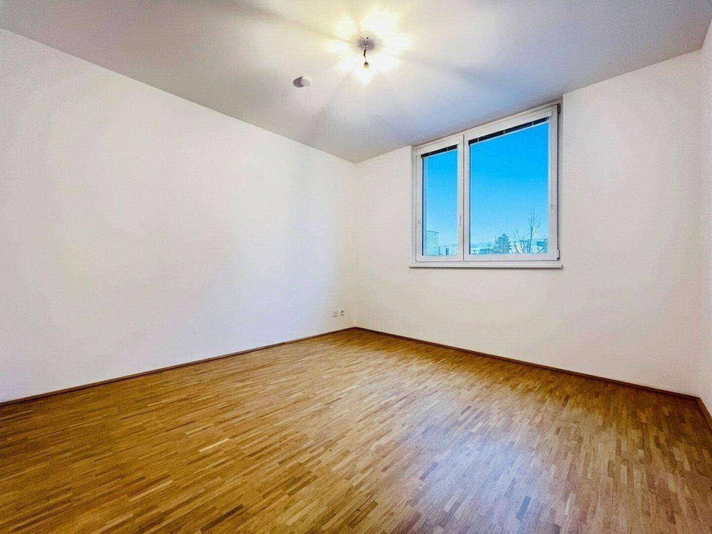 Helles Zimmer mit Parkettboden und einem großen Fenster, das viel Tageslicht hereinlässt.