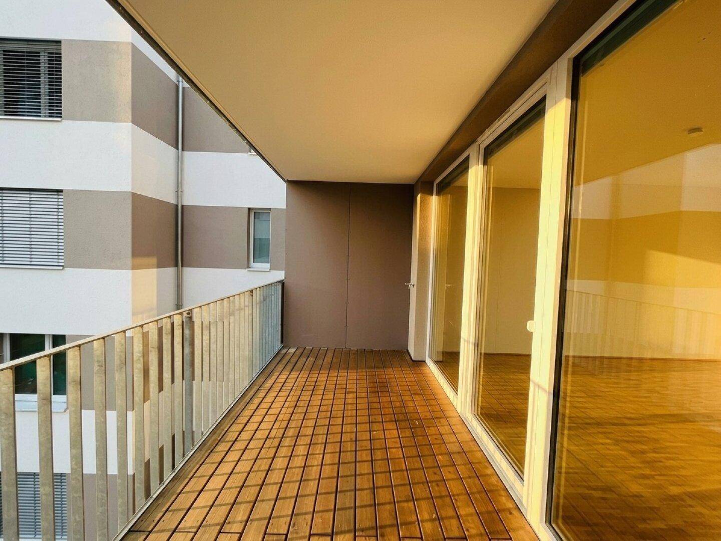 Langer sonniger Balkon mit Holzfliesen und moderner Metallbrüstung, zugänglich vom Wohnbereich.